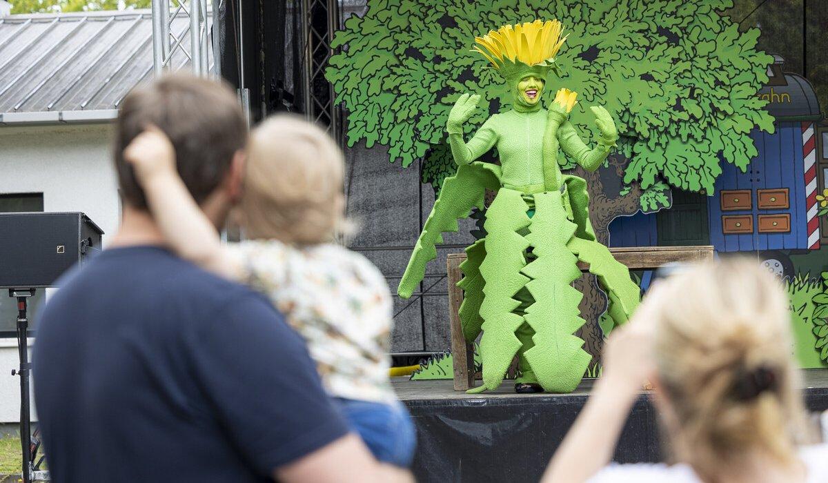 Eine Frau, Vater und Kleinkind schauen einer Person, verkleidet als Baum, auf der Bühne zu