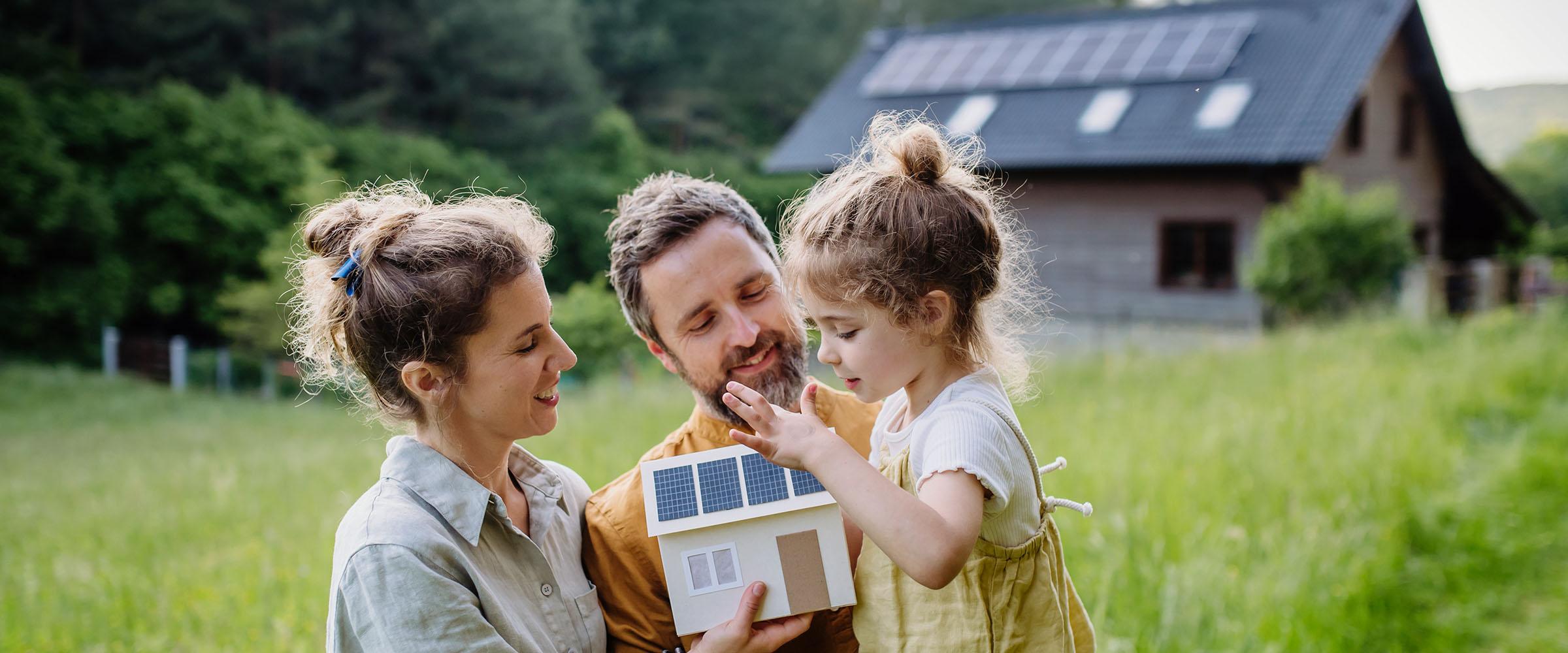 Familie draußen hält ein Modellhaus mit Solarpanels, vor einem Haus mit Solarpanels