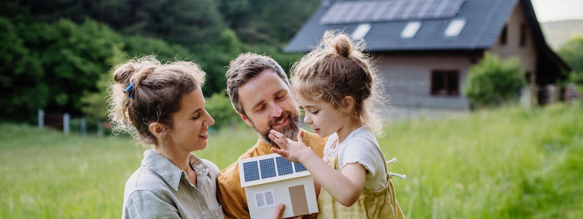 Familie bestehend aus einer Frau, einem Mann und einem kleinem Kind , stehen vor einem Haus, welches mit Solar- Panelen bedeckt ist.